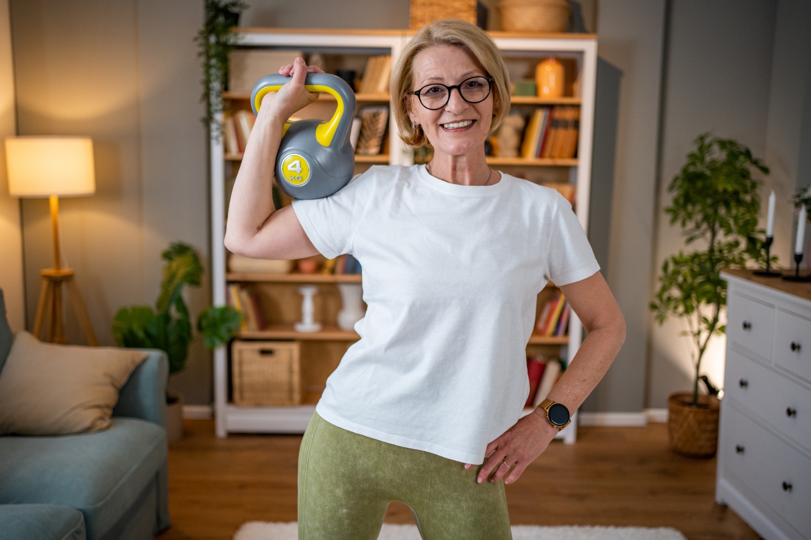 Make 2026 the Year You Finally Stick With Strength Training (Without Relying on Motivation) woman in glasses wearing white shirt lifting weights on her sofa room